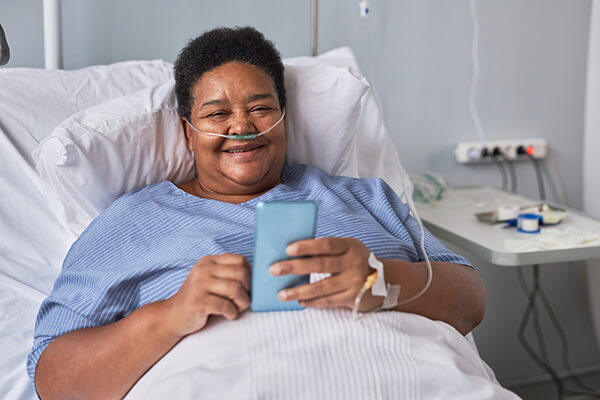 A smiling woman is in a hospital bed and is propped up by pillows. She has breathing tubes in her nose and an IV in her hand. She is using her cell phone to order food.