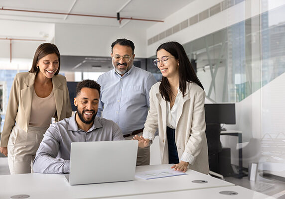 Business team of happy diverse colleagues gathering at laptop computer, watching online content, smiling, laughing. African developer presenting application