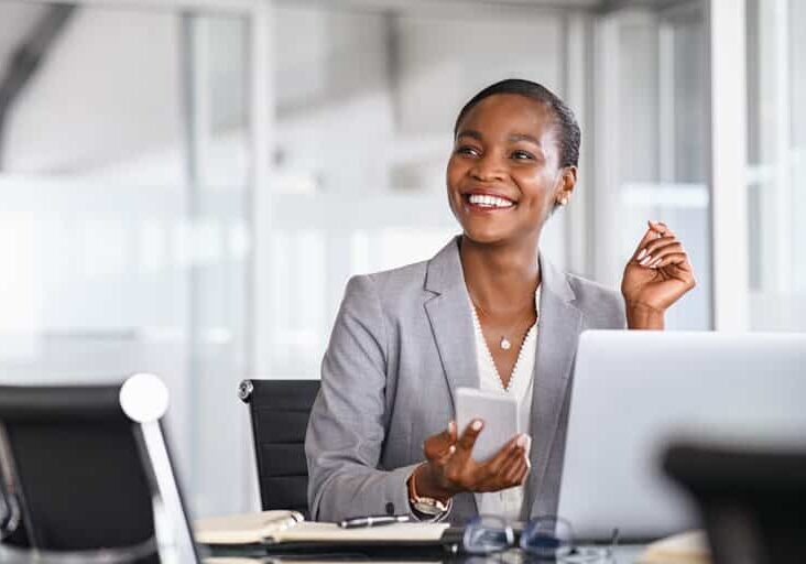 African black business woman using smartphone while working on laptop at office. Smiling mature african american businesswoman lookingup at copy space while working on phone. Successful woman entrepreneur think about new business ideas.