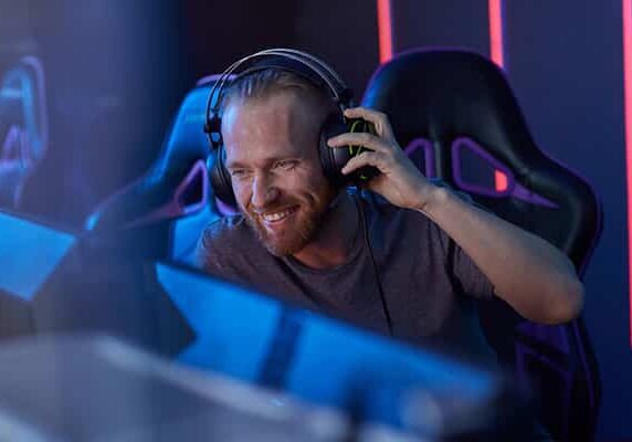 Happy bearded young gamer in headphones looking at computer monitor and laughing while sitting in dark room