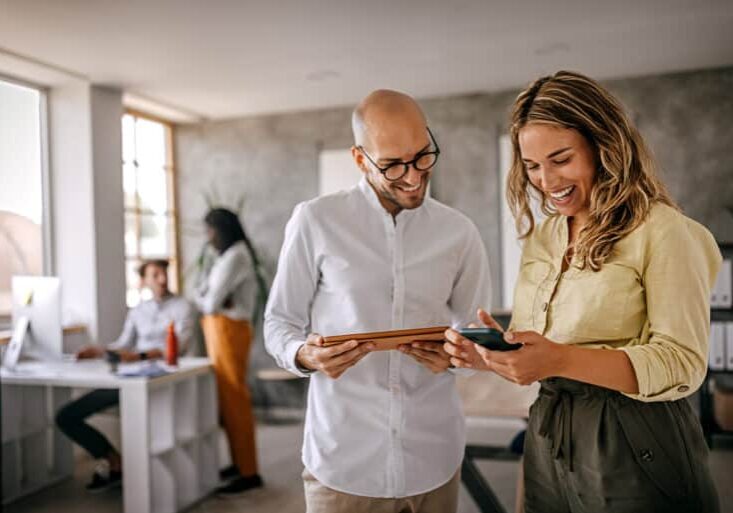 Cheerful and smiling young successful female businesswoman standing with colleague looking at smartphone in modern office and coworking space