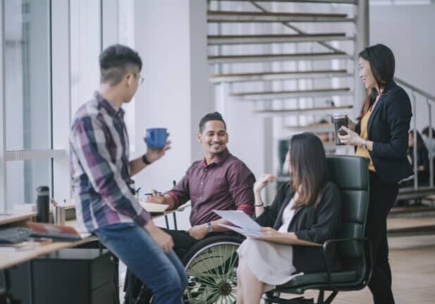 Indian white collar male worker in wheelchair having cheerful discussion leading conversation with colleague in creative office workstation beside window