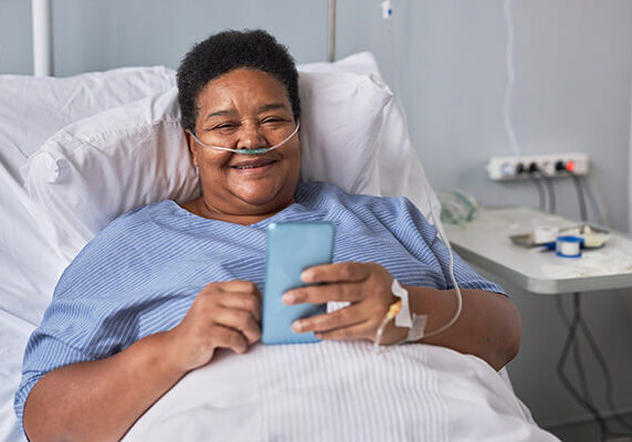 A smiling woman is in a hospital bed and is propped up by pillows. She has breathing tubes in her nose and an IV in her hand. She is using her cell phone to order food.